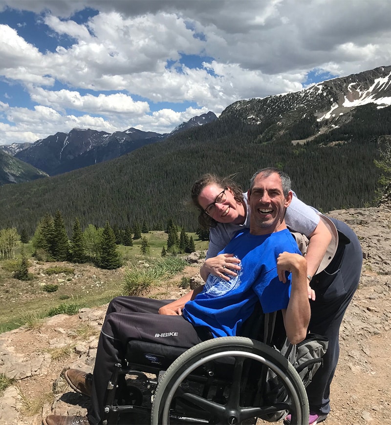 Barton and Megan Cutter, a white male and female, at Molas Pass, Colorado. Barton is in a manual wheelchair wearing a blue four-corners t-shirt. Megan is leaning over beside him wearing a gray four-corners t-shirt. Behind them are several tall pine trees and the San Juan mountains partially covered in snow. A few other tourist are seen on the walkway to a lookout point. Photo taken June, 2023. 