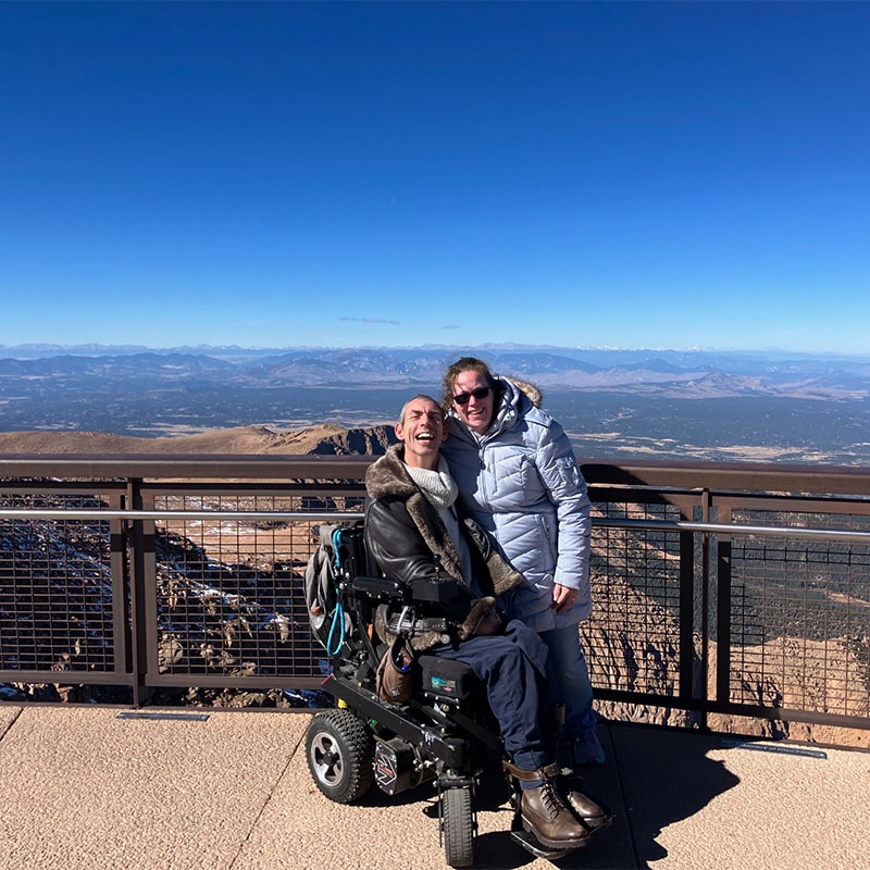 Barton and Megan Cutter, a white male and female, at the summit of Pike’s Peak. Colorado. Barton is sitting in his power wheelchair, and is wearing a white sweater and brown leather coat. Megan is standing beside him wearing a blue sweater and light blue parka with hood. A gray backpack is on the back of Barton’s power wheelchair. Behind them is safety railing, and beyond that are the Rocky Mountains north and west of Pike’s Peak. Photo taken October, 2023.