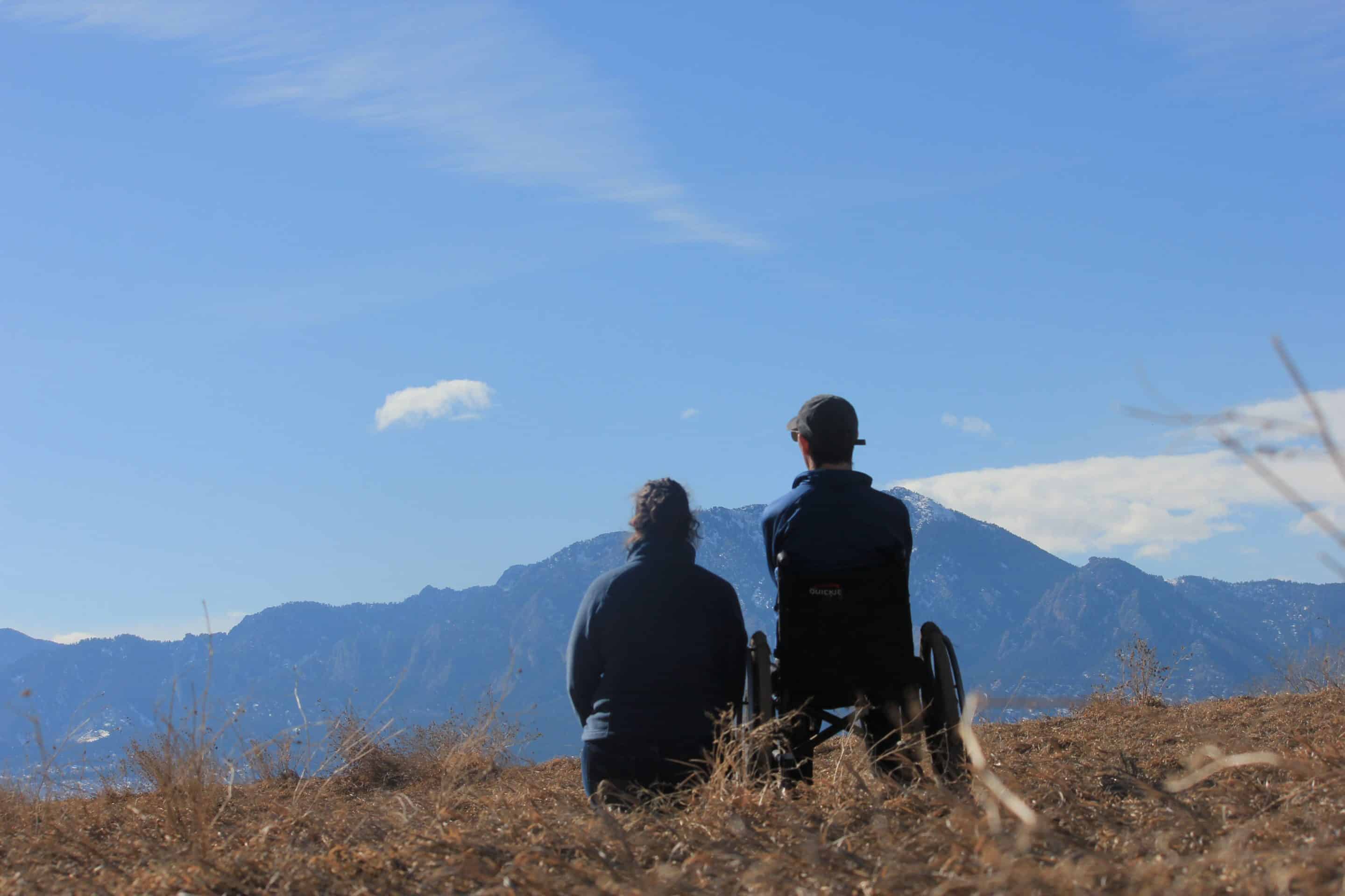 Barton and Megan are smiling in all of the photos below, expressing the joy in their adventures. Barton and Megan Cutter, a white male and female, from behind. Barton is using a power wheelchair, and Megan is turned toward him with her arm around him. They are looking out over Boulder Valley from an elevated lookout at Chautauqua in Boulder, Colorado. A wooden fence is in front of them. Barton is wearing a leather jacket, and Megan’s dark blue hooded coat is draped over the back of his wheelchair. Photo taken February, 2024.