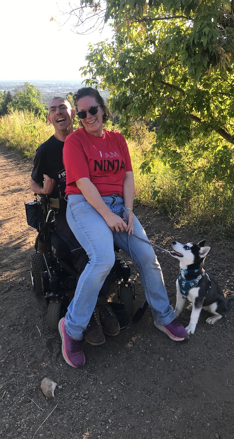 Megan sitting on Barton's lap on a dirt road with their dog