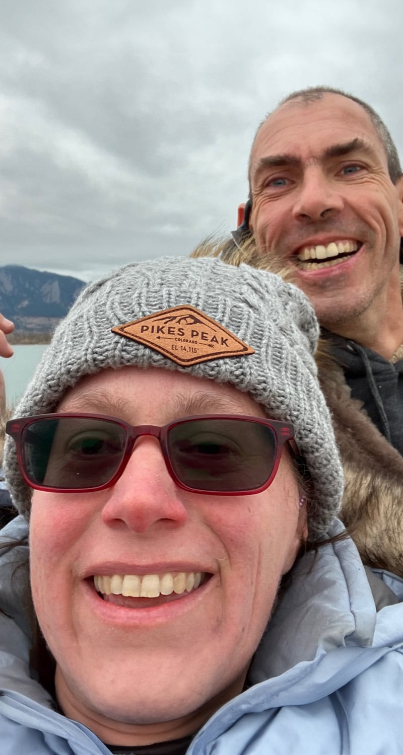 Barton and Megan smiling outside on a cloudy day near the water and mountains.