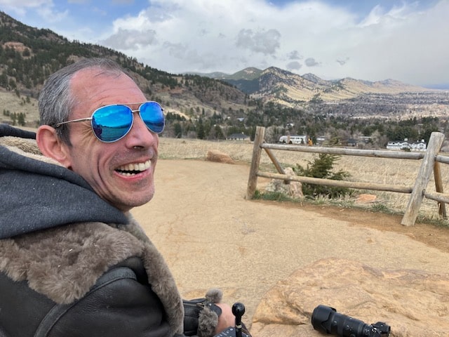 Barton Cutter, a white male, with short gray/black hair and a high forehead. He is wearing sunglasses with blue lenses and a leather jacket. He is looking backwards over his right shoulder at the camera. In front of him is a split wooden fence and mountains in the background. There are clouds with a patch of blue sky above his head. He is at Chautauqua in Boulder, Colorado. Photo taken February, 2024. 