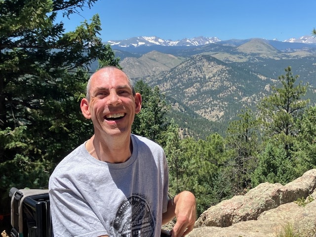 •	Barton Cutter, a white male, with short gray/black hair and a high forehead, is sitting on an outcropping area with granite rock and gravel from the summit of Flagstaff Mountain, behind Chautauqua in Boulder, Colorado. There is one tall pine tree behind him and the Rocky Mountain range to the right of his head. The sky is blue, with no clouds. He is wearing a gray four-corner’s t-shirt. Photo taken August, 2024.