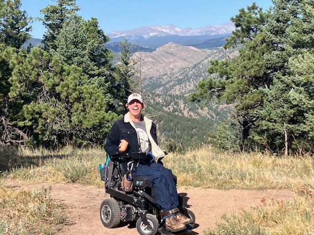 Barton Cutter, a white male, with short gray/black hair and a high forehead, is sitting in his wheelchair facing forward in an outcropping area just before the summit of Flagstaff Mountain, behind Chautauqua in Boulder, Colorado on a dirt trail. There are pine and ponderosa trees on either side of him and the Rocky Mountain range behind his head. Barton is wearing jeans, brown boots, t-shirt and a blue corduroy jacket. He is wearing a ball cap. The sky is blue, with no clouds. Photo taken July, 2024.