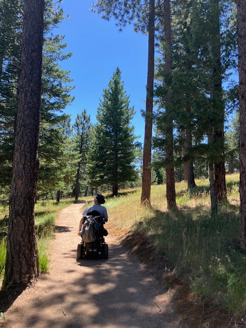 Barton Cutter, a white male, with short gray/black hair and a high forehead, is rolling down a dirt hiking trail path using his power wheelchair going to summit of Flagstaff Mountain (Ute Wheelchair trail), behind Chautauqua in Boulder, Colorado. Barton is wearing a brown brimmed hat. There are tall pine and ponderosa trees on either side of the dirt path. Photo taken August, 2024.