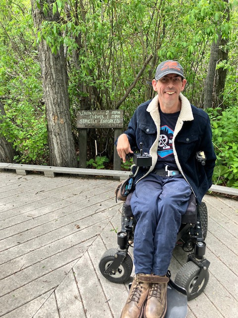 Barton Cutter, a white male, wearing jeans, t-shirt, and a blue corduroy jacket and baseball cap, is sitting in his power wheelchair on a wooden walkway in the middle of dense woods. There is a sign behind him that reads “Whoaw 2mph, curves 1 mph, strickly enforced in white paint and “WOW vigilantes” underneath in gray. He is at Wilderness on Wheels, in Grant, Colorado. Photo taken June 2024. 