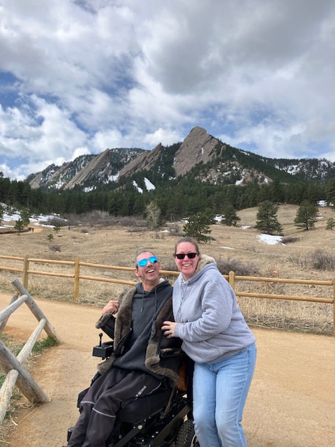 Barton and Megan Cutter, a white male and female, faceing forward. Barton is using a power wheelchair, and Megan has with her arm around him, slightly kneeling. Behind them are the Flatiron rock mountains from an elevated lookout at Chautauqua in Boulder, Colorado. A wooden fence is to the left of Barton and also behind them. Barton is wearing a leather jacket, and Megan is wearing jeans and a gray sweatshirt. Photo taken February, 2024. 