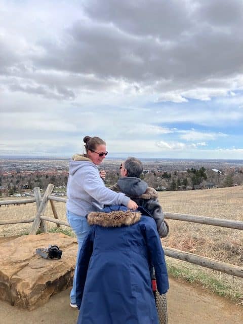 Barton and Megan Cutter, a white male and female, from behind. Barton is using a power wheelchair, and Megan is turned toward him with her arm around him. They are looking out over Boulder Valley from an elevated lookout at Chautauqua in Boulder, Colorado. A wooden fence is in front of them. Barton is wearing a leather jacket, and Megan’s dark blue hooded coat is draped over the back of his wheelchair. Photo taken February, 2024. 