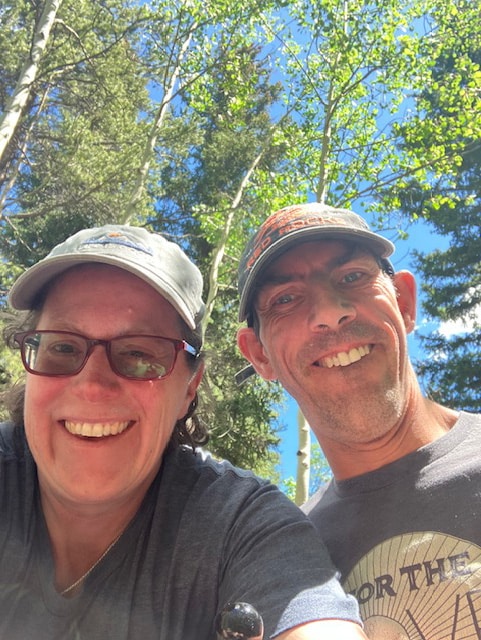 Barton and Megan Cutter, a white male and white female, face directly into the camera. They both are wearing ball caps and t-shirts, and Megan is wearing glasses. The are on a wooden walkway in the middle of dense woods behind them. Barton is sitting his power wheelchair. They are at Wilderness on Wheels, in Grant, Colorado. Photo taken June 2024.