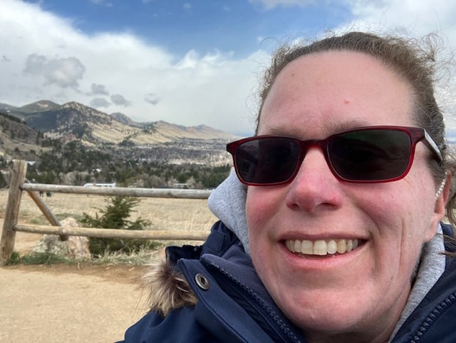 Megan Cutter, a white female, is looking straight ahead. She is wearing sunglasses, and a dark blue winter coat. There is a split wooden fence and mountains in the background. There are clouds with a patch of blue sky above her. She is at Chautauqua in Boulder, Colorado. Photo taken February, 2024. 