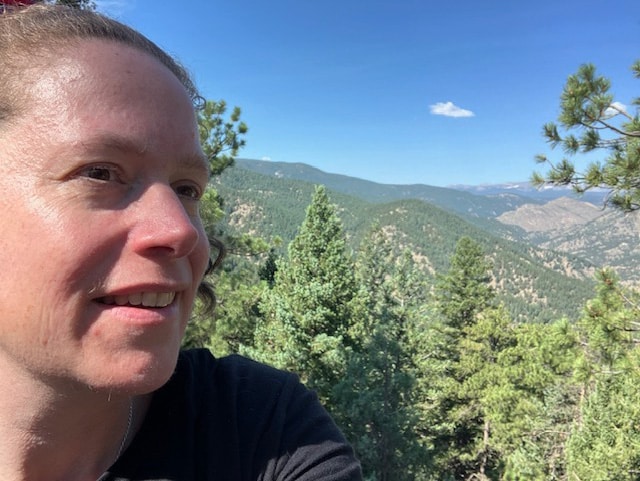 •	Megan Cutter, a white female in profile, is overlooking a wooded area from the summit of Flagstaff Mountain, behind Chautauqua in Boulder, Colorado. The sky is blue with one small cloud. She is wearing a black t-shirt. Photo taken August, 2024.