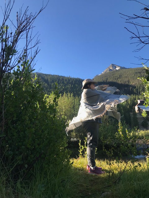 •	Megan Cutter, a white female, is standing in an open grassy area beside a river. She is wearing blue jeans and is wrapped in a gray patterned shawl, which is flaring out at the bottom from being flung around her. In the background is a pine forest and a mountain peak with some slight snow covering. She is camping with Barton in Silverton, Colorado. Photo taken July, 2023. 